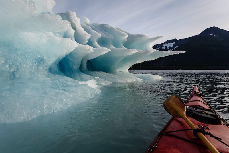 Paddler lashes paddle to kayak to pause journey to enjoy the splendor of the shapes of the melting Iceberg in summertime Alaska's Kenai Fiords National Park