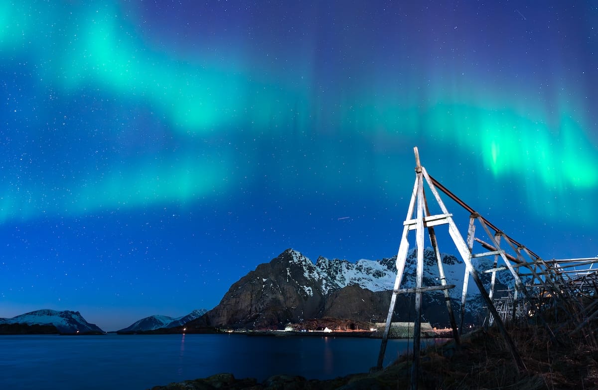 Northern lights glowing green and turquoise over a fjord, snow-dusted mountains, and a wooden stockfish drying rack in the Lofoten Islands, Norway