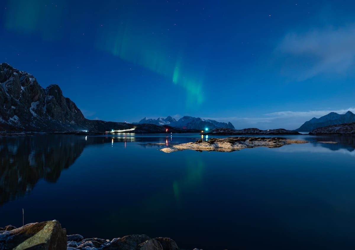 Faint northern lights over a calm fjord reflecting the night sky, with rocky shoreline and snowy mountains in the distance, seen from Svinøya Rorbuer in Svolvær, Lofoten
