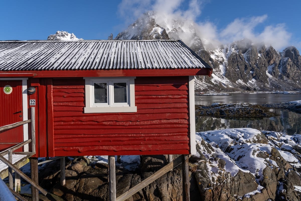 Things to do in Svolvær travel guide (Svinøya Rorbuer) Red wooden rorbu (fisherman’s cabin) on stilts over the rocks and water in Svolvær, Lofoten, with snow on its roof and misty snow-capped mountains behind