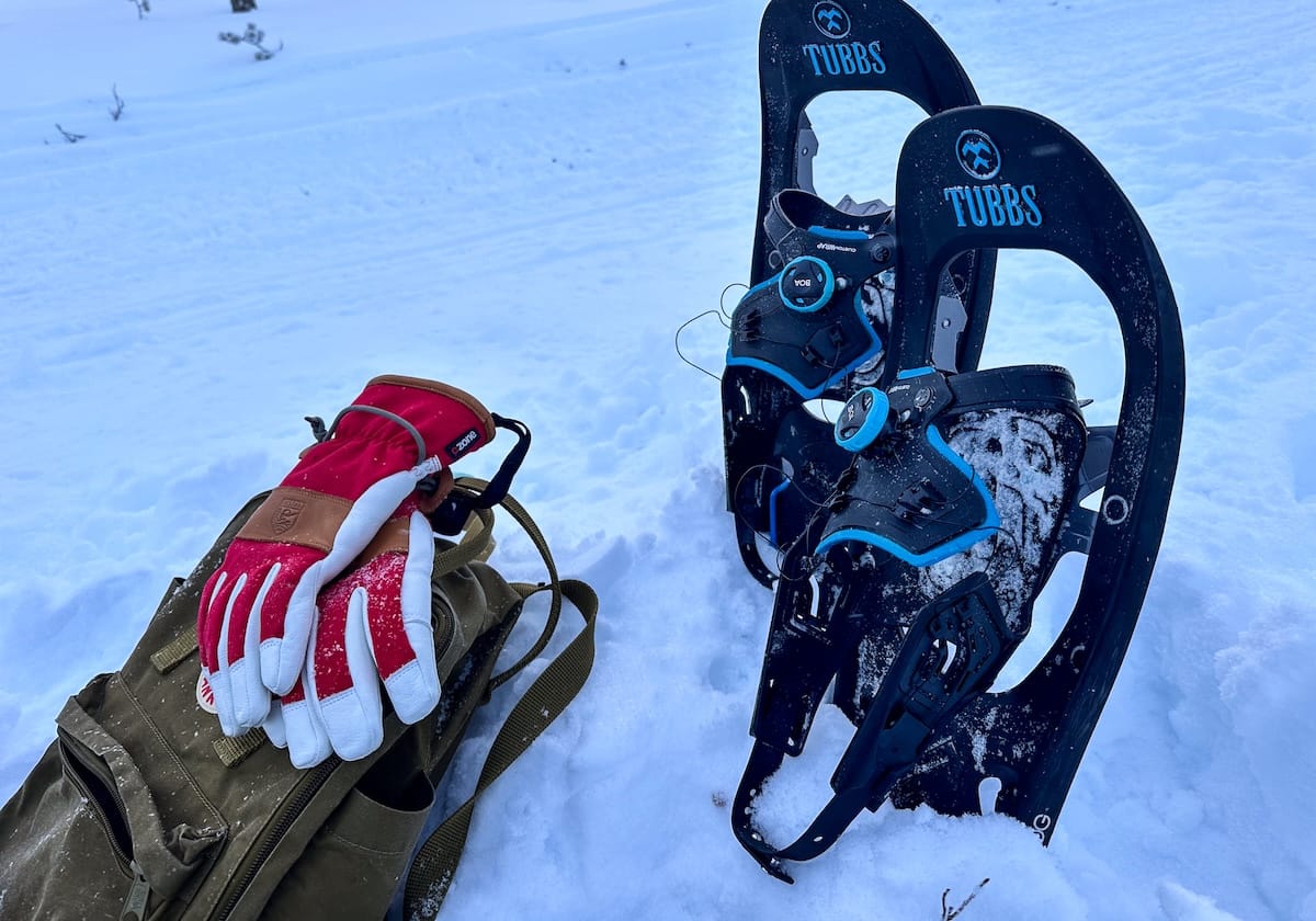 Pair of Tubbs snowshoes standing upright in deep snow next to a pair of red and white winter gloves resting on an olive green backpack