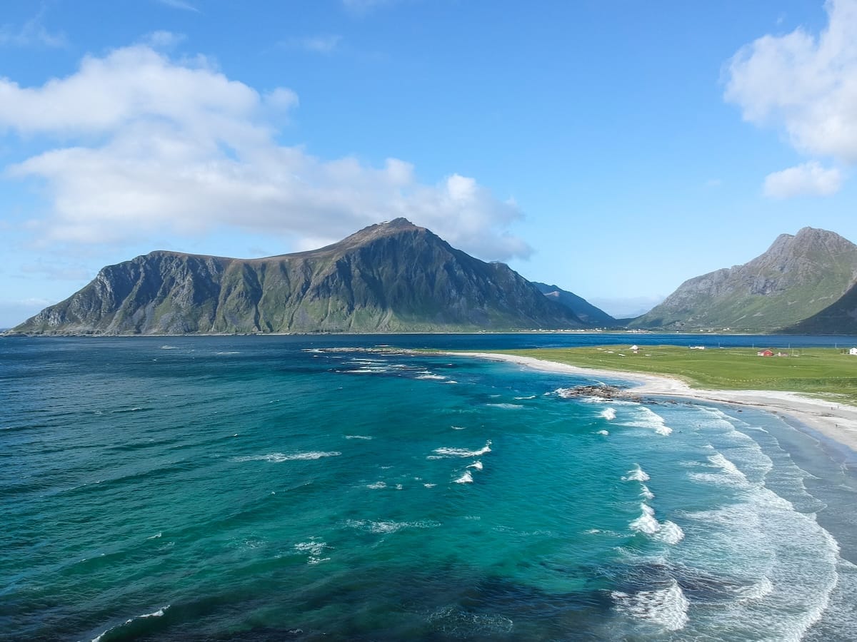 Turquoise waves rolling onto a white sand beach in the Lofoten Islands, with green mountains and a few small houses along the shore under a blue summer sky