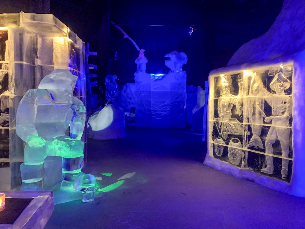 Illuminated ice sculptures at Magic Ice Lofoten in Svolvær, including a seated figure in a hood and a carved relief of fishermen with fish and baskets, lit in blue and green