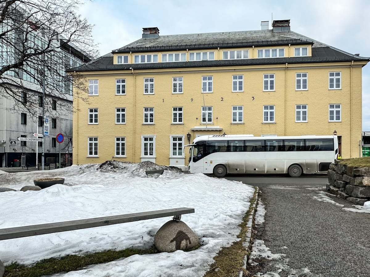 Yellow historic building housing the Lofoten War Memorial Museum (Lofoten Krigsminnemuseum) in Svolvær, with a white tour bus parked in front and patches of snow in the foreground