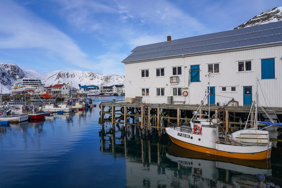 Photo of Honningsvåg, Norway's harbor area and boats during early April. There is snow on the mountains in the background and a partially blue sky.