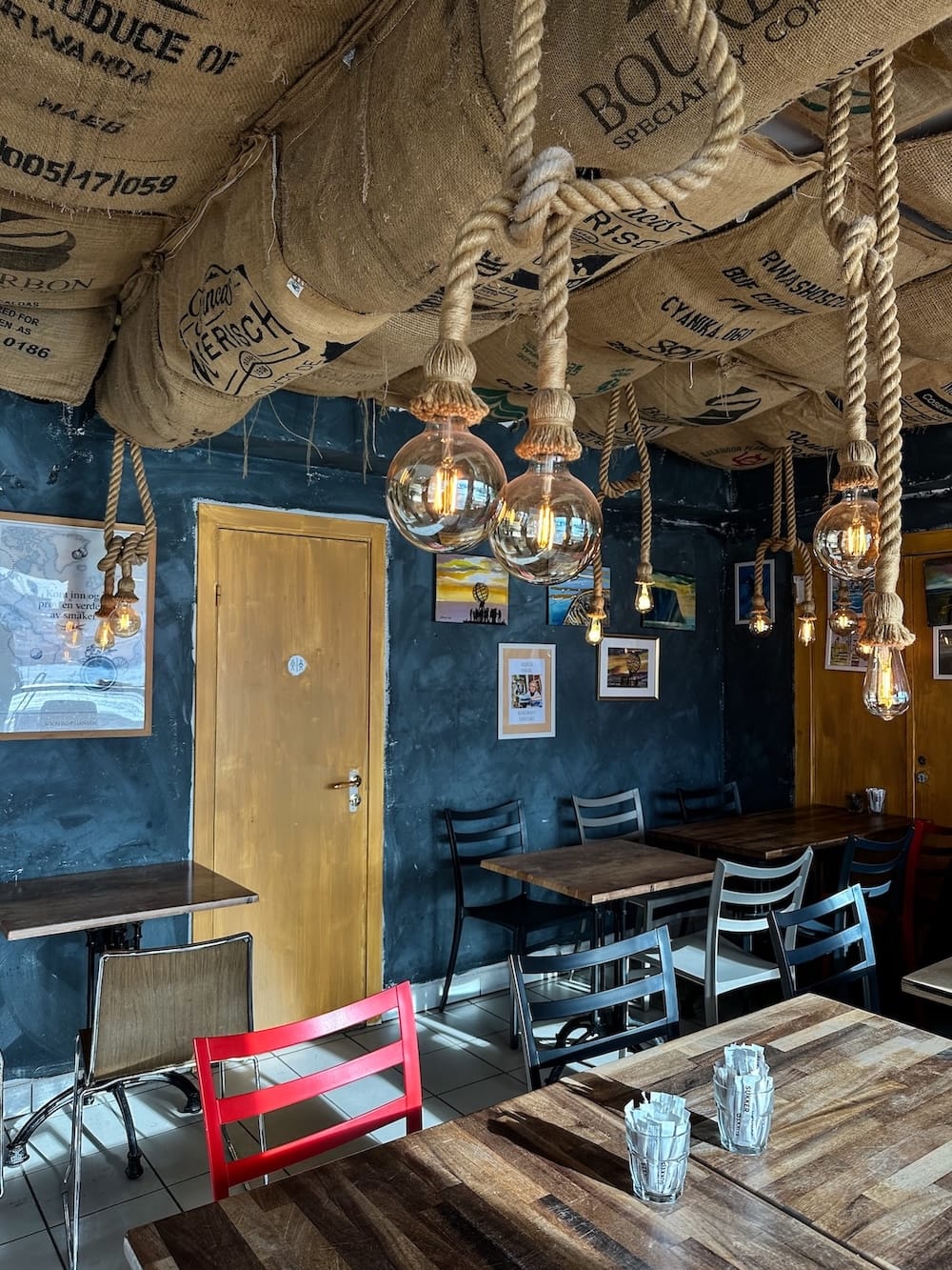 This is a cafe called Honni Bakes in Honningsvåg, Norway. The photo has coffee bean bags on the ceiling with ropes hanging down and colorful chairs.