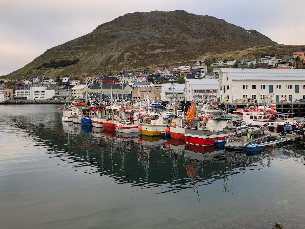 The harbor of Honningsvåg, Norway during June - there is a mountain in the background with boats in the foreground. It is brown and rocky without a lot of color except the boats.