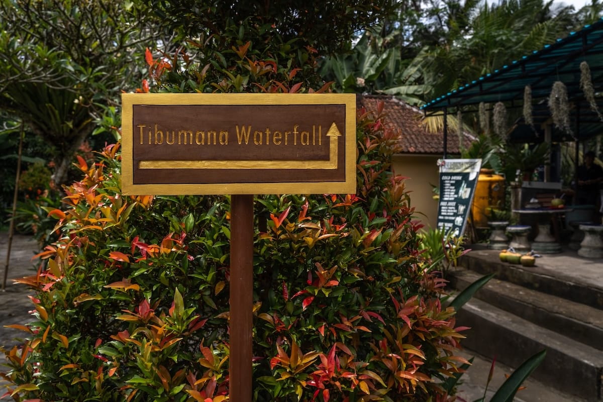 This is a sign that says 'Tibumana Waterfall' on it and it is situated around orange flowers