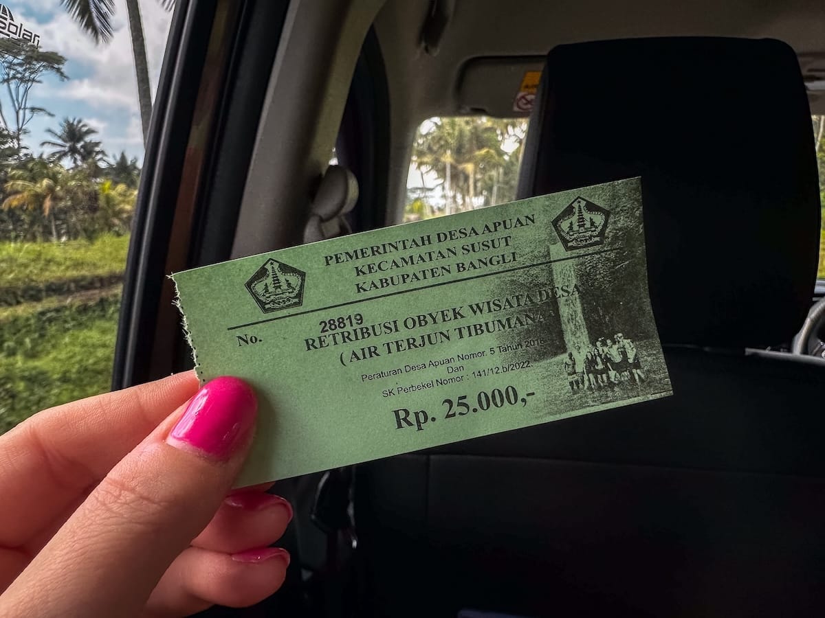 This is a photo of a ticket for a waterfall in Bali - it is green in a hand of a woman and shows the price of 25000 IDR