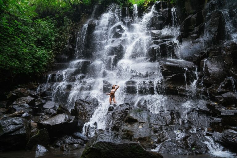 A Complete Guide to Visiting Kanto Lampo Waterfall in Bali This is a photo of a girl standing in front of a big waterfall on a rock in Bali