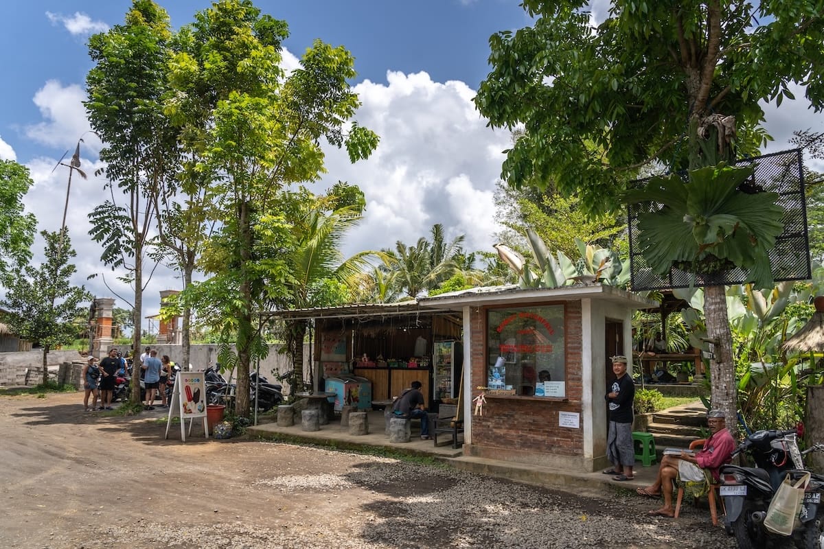 This is a ticket counter for a waterfall called Suwat in Bali. There are a few people sitting outside of it and there is no one in line.