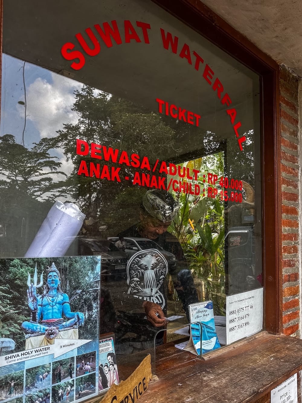 This is a ticket counter for Suwat Waterfall in Bali - it has red writing on the window and a man standing behind the glass window