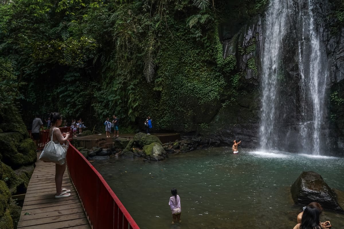 This is a red bridge that is overtop of a natural pool at Ulu Petanu Waterfall. You can see the waterfall and some people in it.