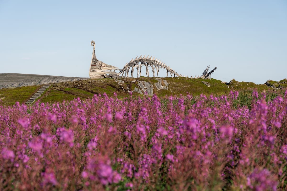 A wooden statue on top of a hill with purple flowers. The statue is in the shape of both a whale and a viking ship.