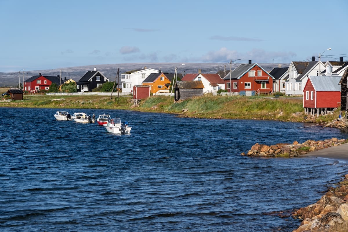 The village of Skallelv, Norway - it is on the road to Vardø and this photo has a bunch of yellow, red, and white homes on a small harbor with a few small fishing boats. The sky is blue and sunny.