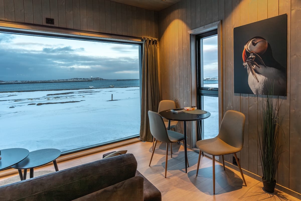 This is Varanger View lodging. It is a small cabin with a huge window to see out of (and the fjord in the background). There is a small table with three chairs, a photo of a puffin on the wall, and a couch.