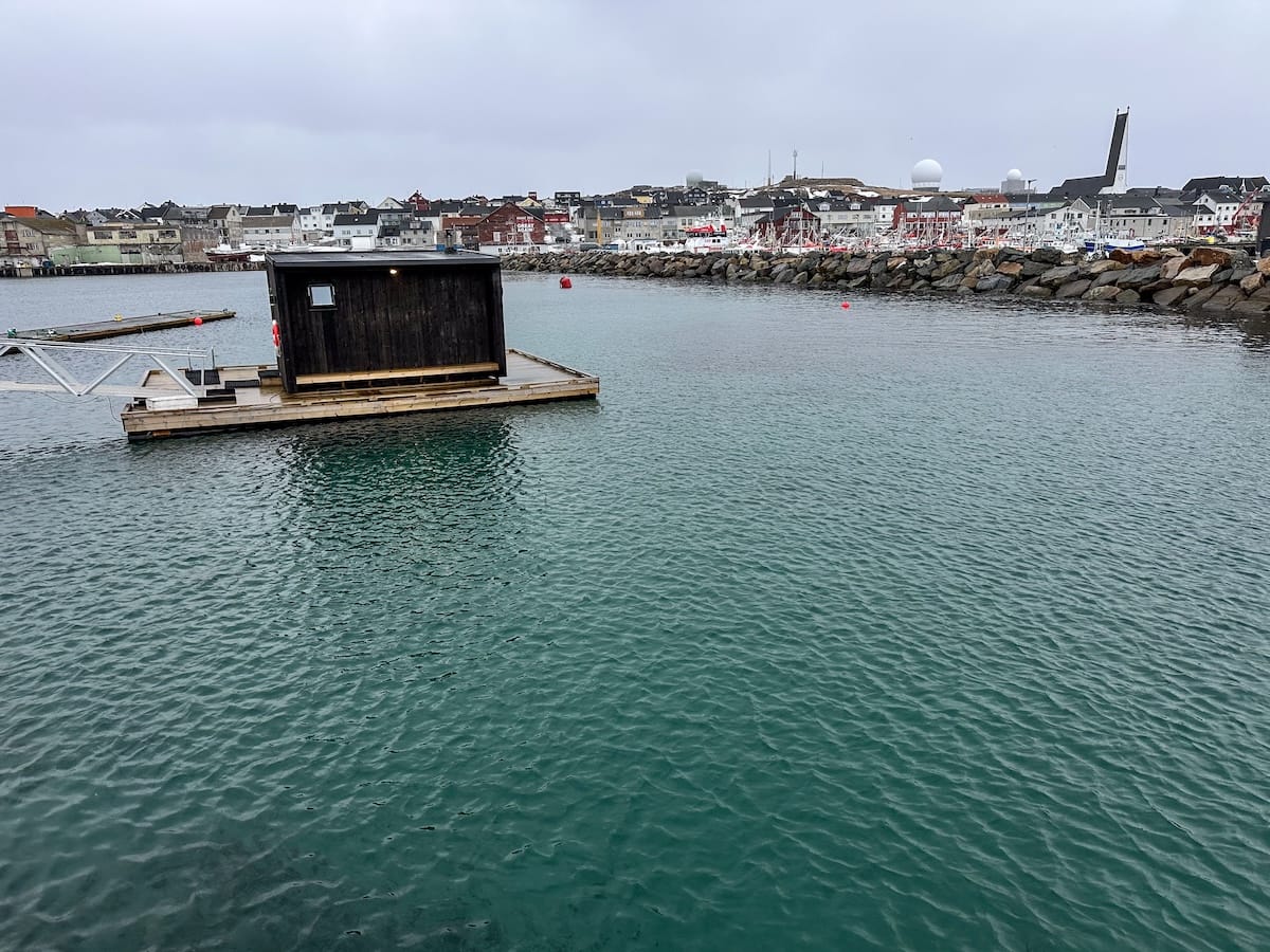 A black wooden sauna that is floating in a harbor in Vardø. There is a pier going down to it and the water is a tealish green.