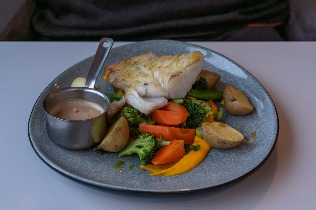 A plate of fresh cod, a rhubarb sauce in a side pan, carrots, broccoli, potatoes, and an orange colored puree.