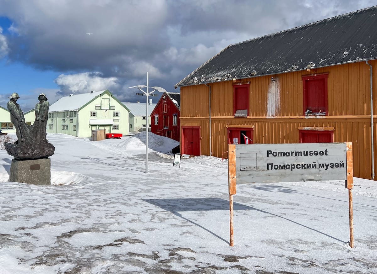 A Pomor history museum in Vardø Norway - the museum is yellow with a sign in front (in both Norwegian and Russian) with a statue of two men to the left. There is snow on the ground
