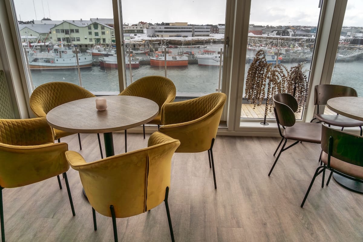 Yellow chairs and a circular table in front of a large window and terrace in front of a harbor and boats in Vardø, Norway