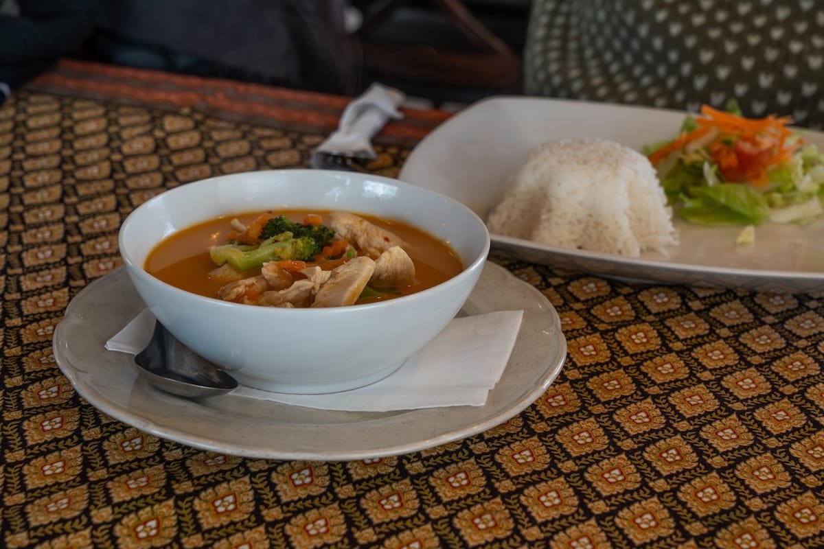 A photo of a red Thai curry in front of a plate of rice on a colorful tablecloth.