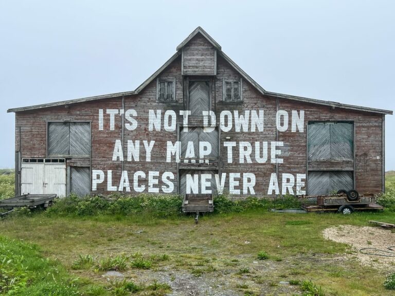 One day in Vardø itinerary | What to do with 24 hours in Vardø, Norway This is a photo of an old grey wooden building with the words 'It's Not Down on Any Map, True Places Never Are' written largely across it. The sky is grey.