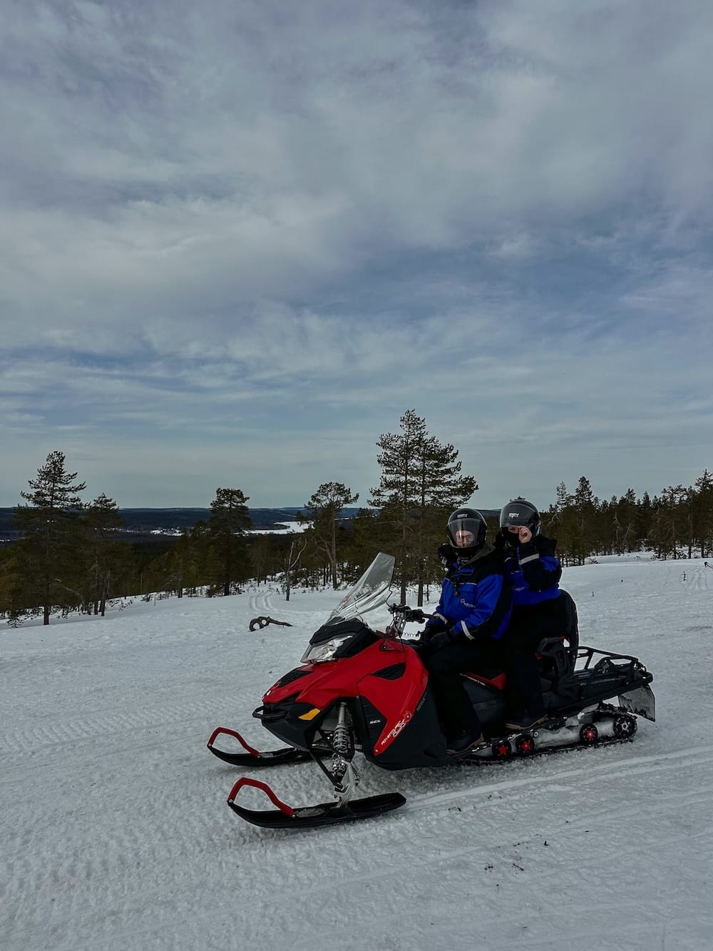 Photo: this is me (a woman) and my partner (a man) in our winter suits on top of a snowmobile in a snowy, Finnish landscape