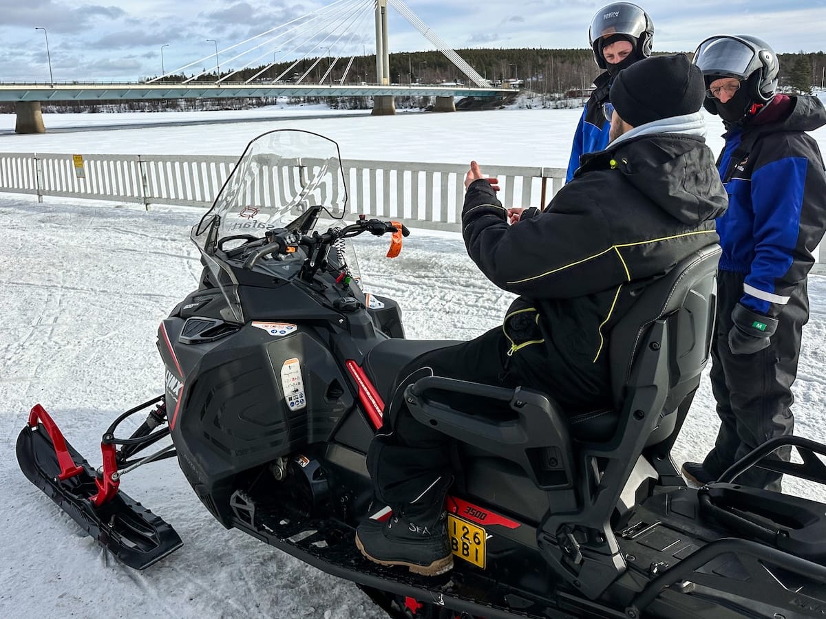 Photo: Getting briefed about how to operate a snowmobile by the frozen river in Rovaniemi, Finland. This shows two tour participants and a bridge in the background.