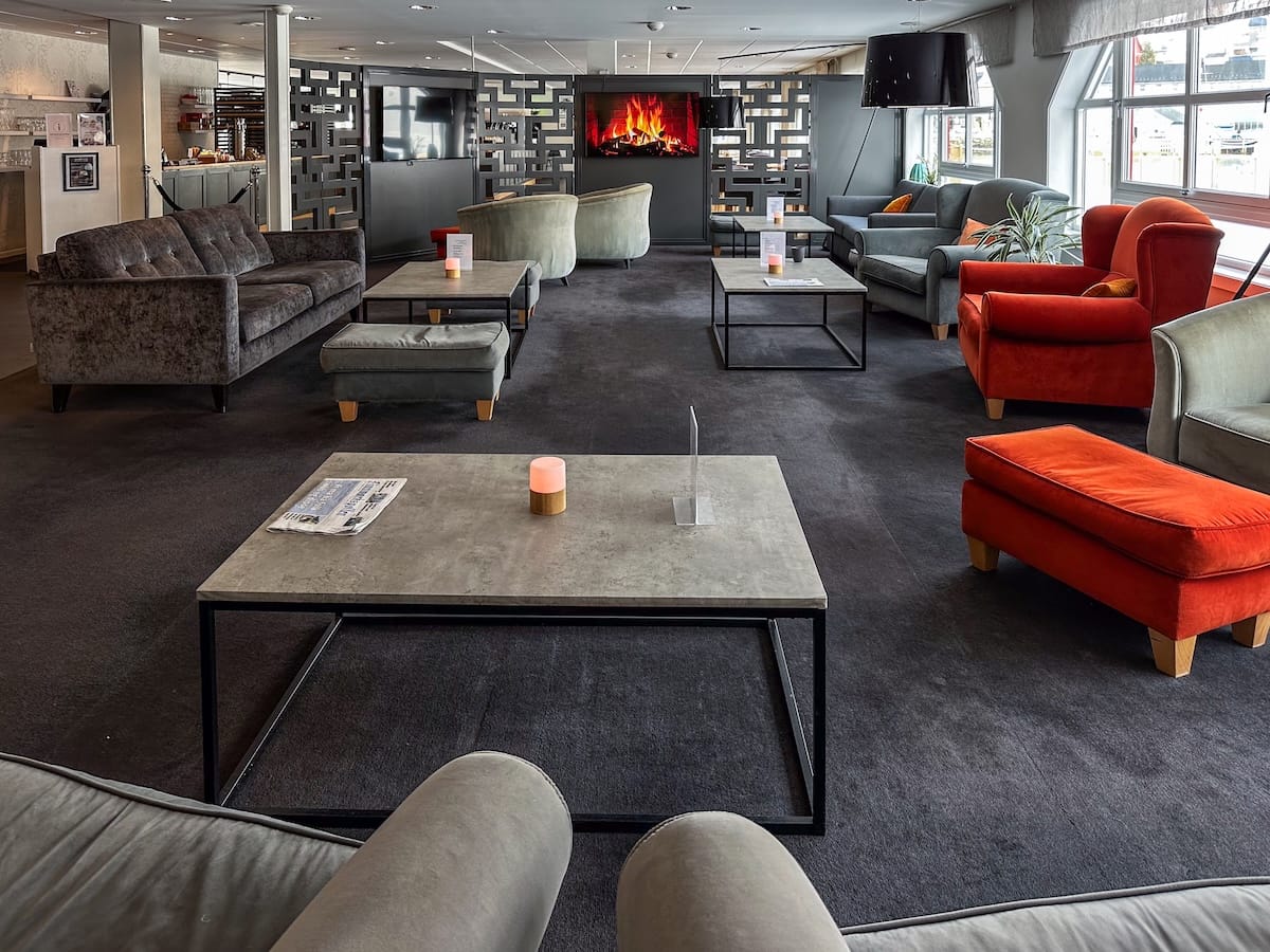 The lobby at Scandic Bryggen in Honningsvåg, Norway.
This photo is a hotel lobby with grey and red furniture, tables, and a fake fireplace.