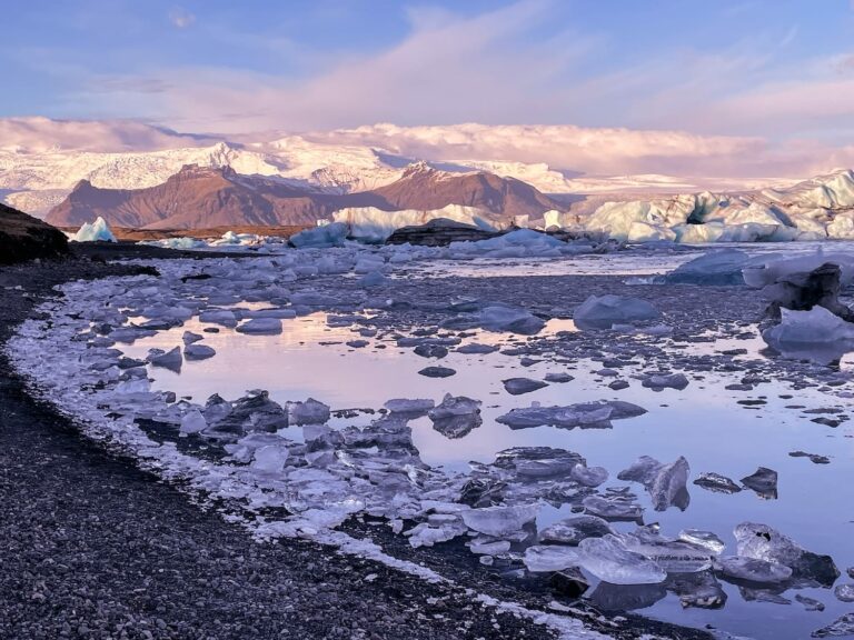 How to Visit Jökulsárlón Glacier Lagoon in Iceland This is a photo of a blue/purple sky in front of the glacier lagoon in iceland. Lots of icebergs scattered in the water