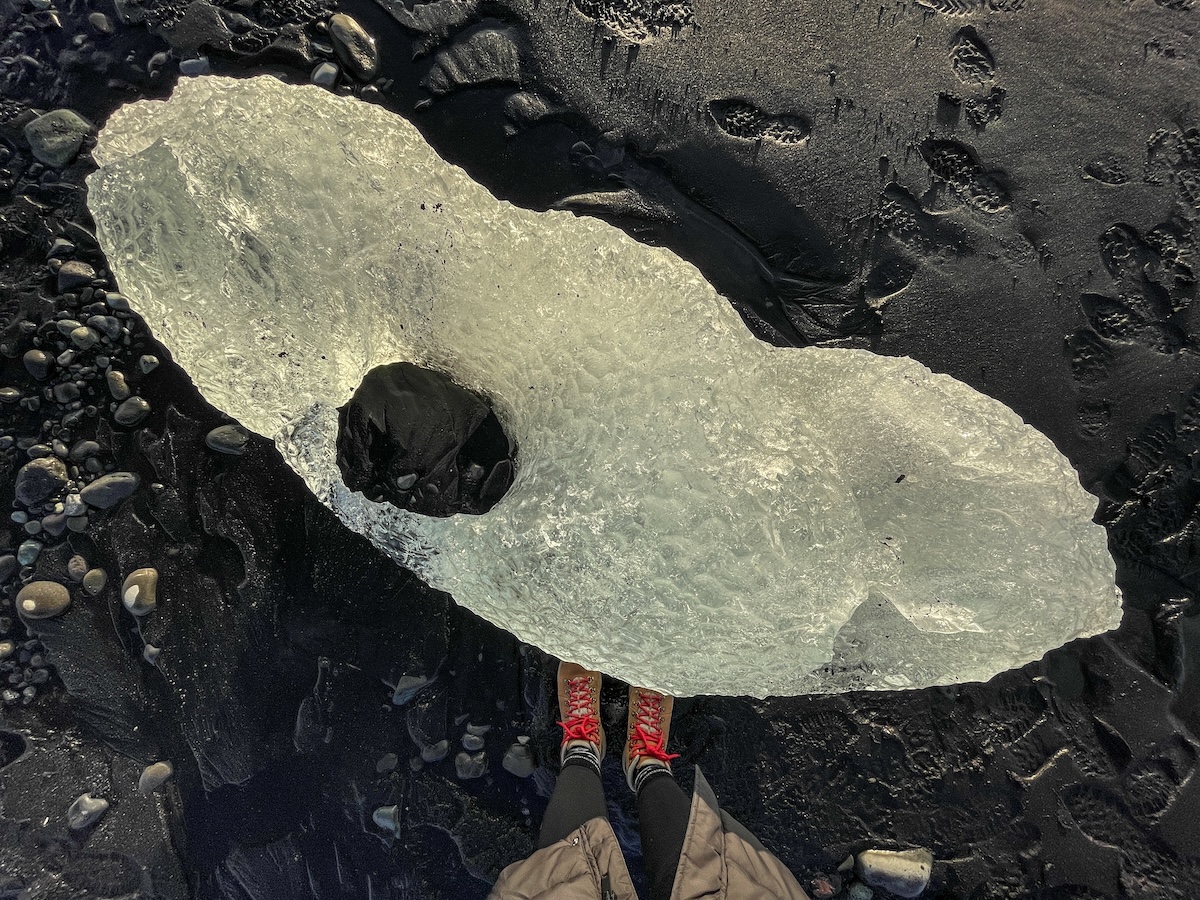 A girl in hiking boots (only her feet are shown) with red laces standing in front of a small iceberg on a black sand beach in Iceland