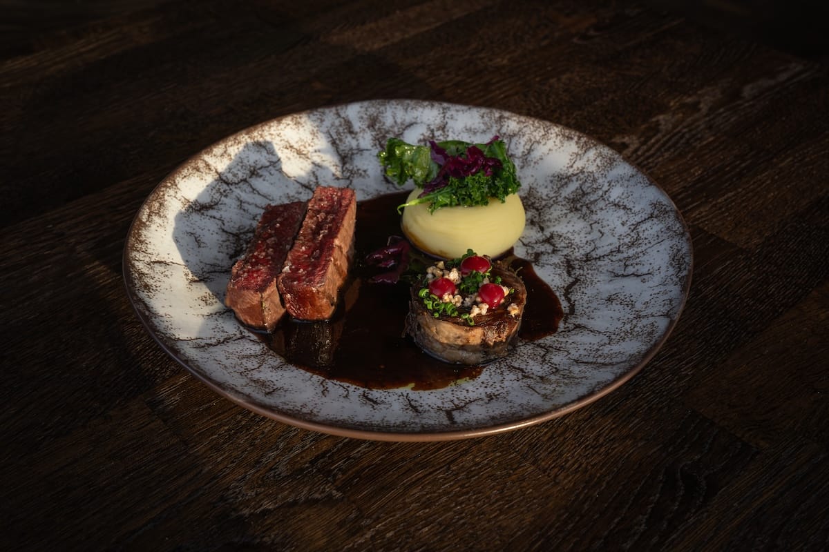 A plate with a reindeer sirloin, a reindeer entrecote wrapped with lingonberry on top, mashed potatoes topped with kale, and a game sauce.