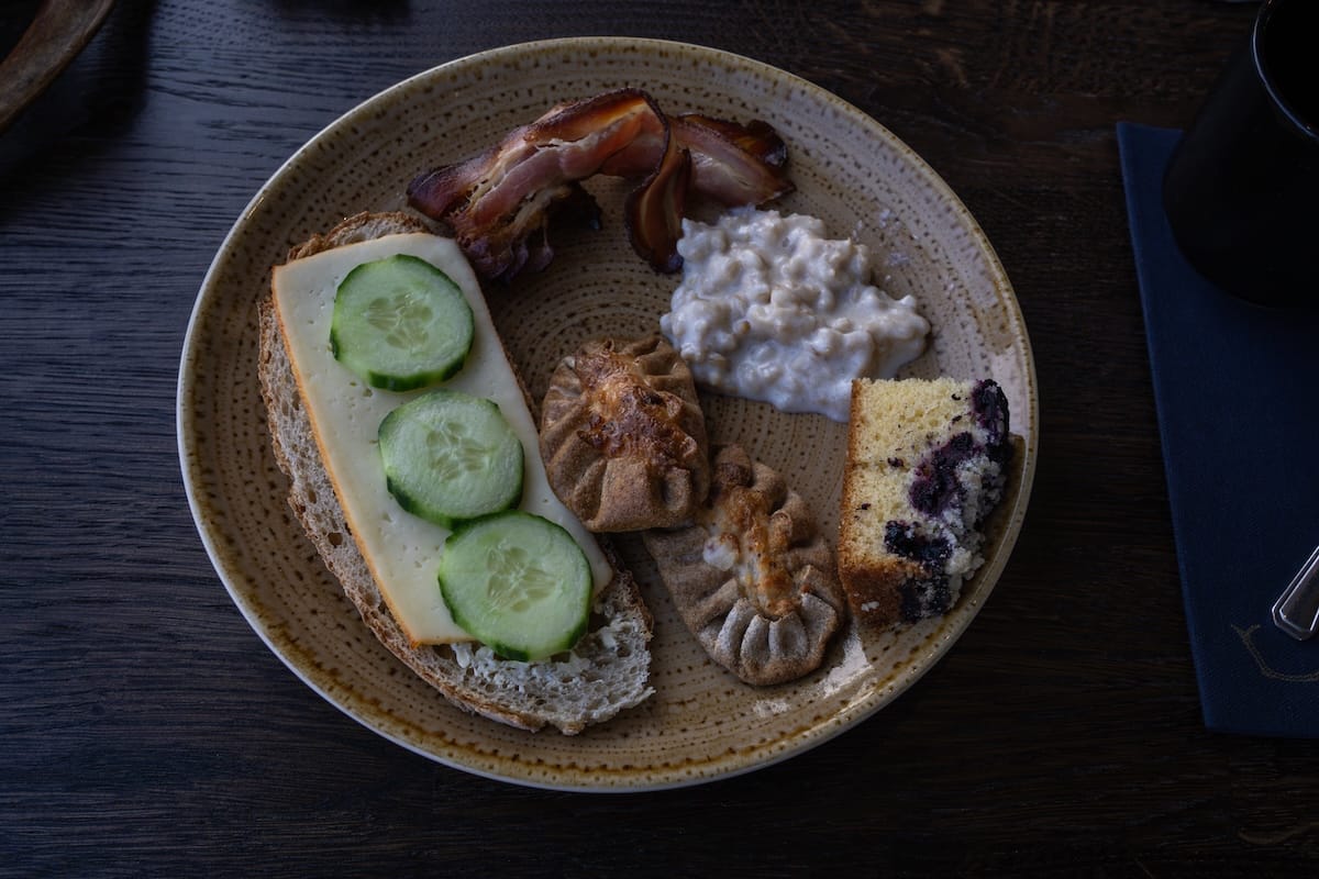 A plate of food from breakfast: bread topped with cheese and cucumbers, a karelia pie, a blueberry cake, porridge, and bacon