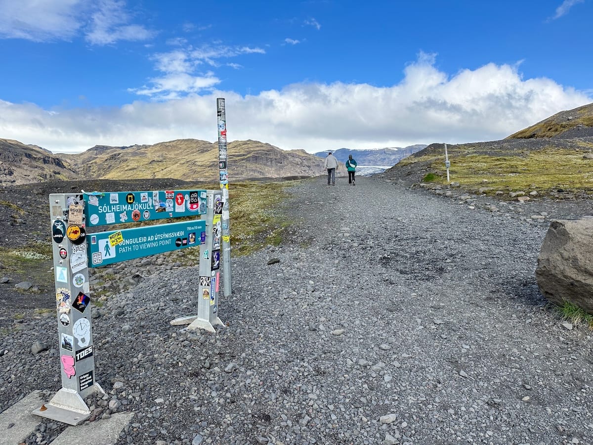 This is a photo leading up to Sólheimajökull Glacier in Iceland along the South Coast. There is a sign with stickers all over it and a gravel path leading to the glacier.