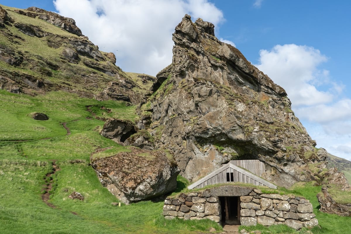 Rútshellir on the South Coast
This is a photo of a sharp large rock with a small cave structure (man-made) underneath. The grass is green and the skies are blue.