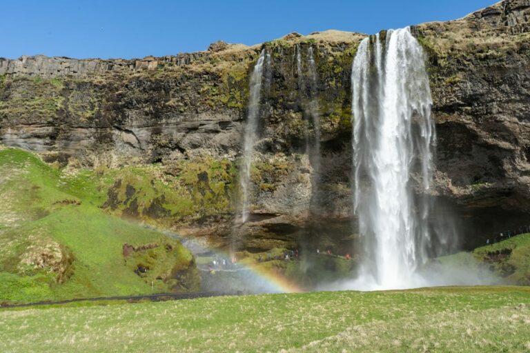 Visiting Iceland in May | A May in Iceland travel guide This is a photo of Seljalandsfoss in Iceland with a rainbow at the bottom of the waterfall and blue skies and green grass.