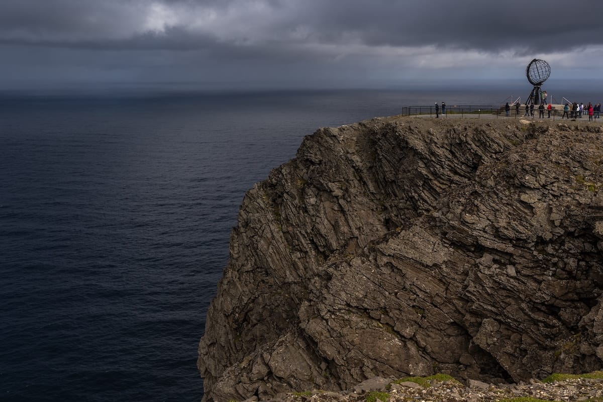 Where to Stay at the North Cape in Norway (+ Honningsvåg)
a photo of a tall cliff with a globe on top of it with people around it - there are stormy skies over the sea in the distance