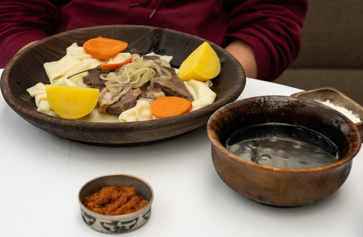 A plate of beshbarmak on a white table with a side of sorpa and chili