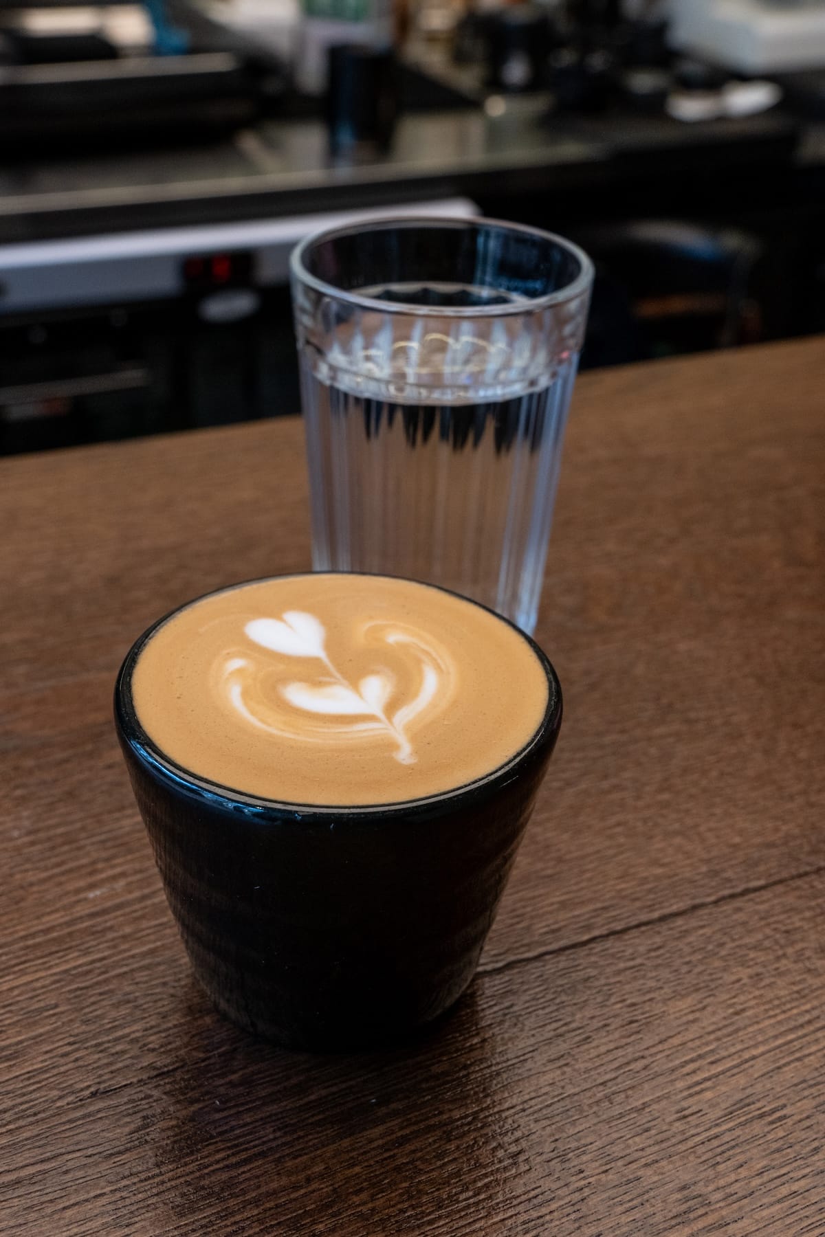 a coffee in a black cup on a counter (with a glass of water behind)