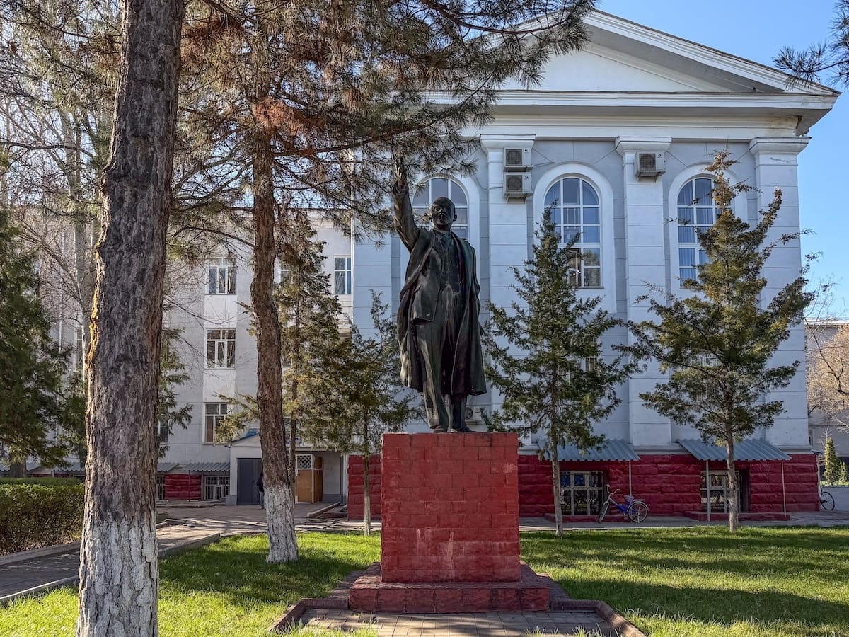 a statue of lenin at a university - red base and blue building behind