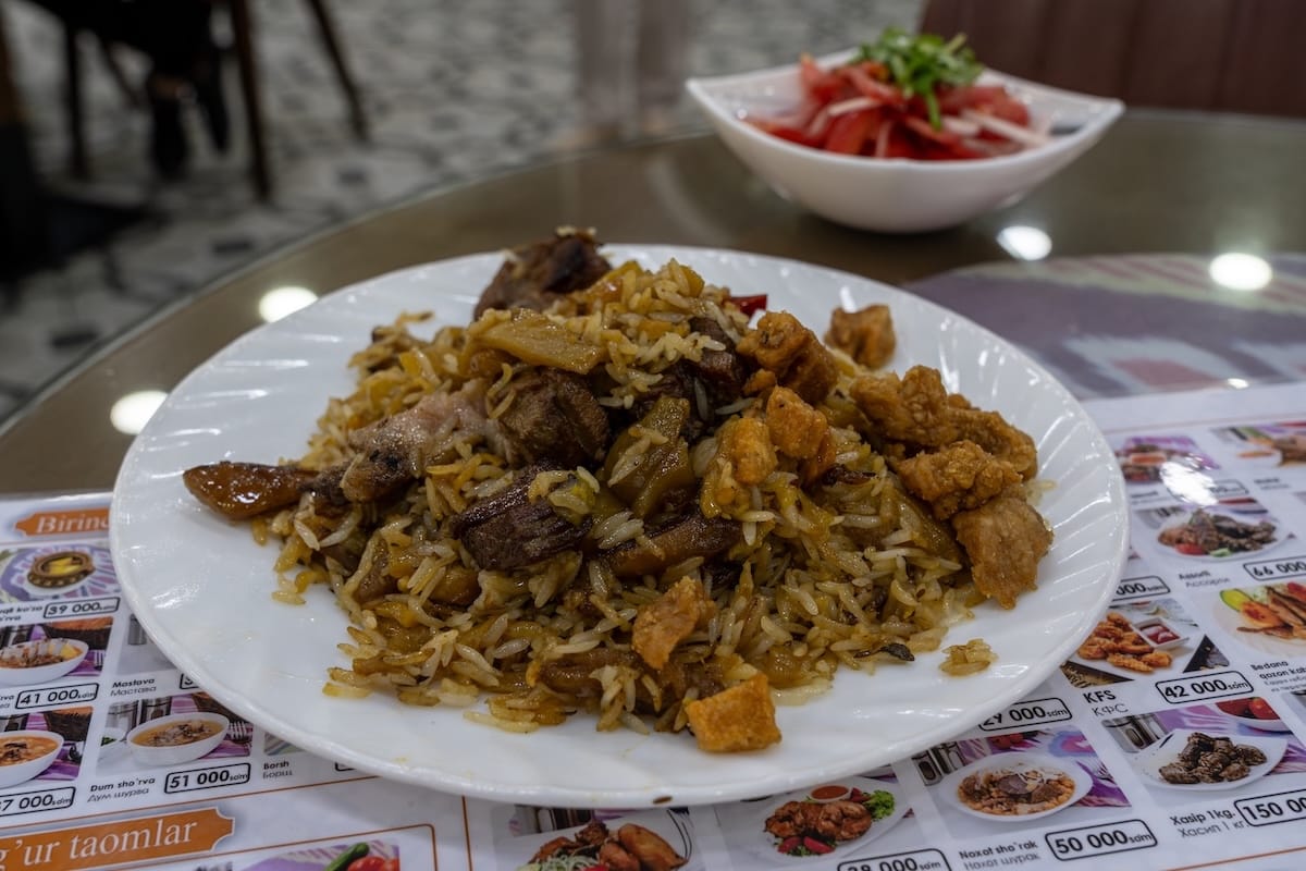 a plate of plov on a table with crispy lamb fat on top of it and a bowl of tomatoes behind it