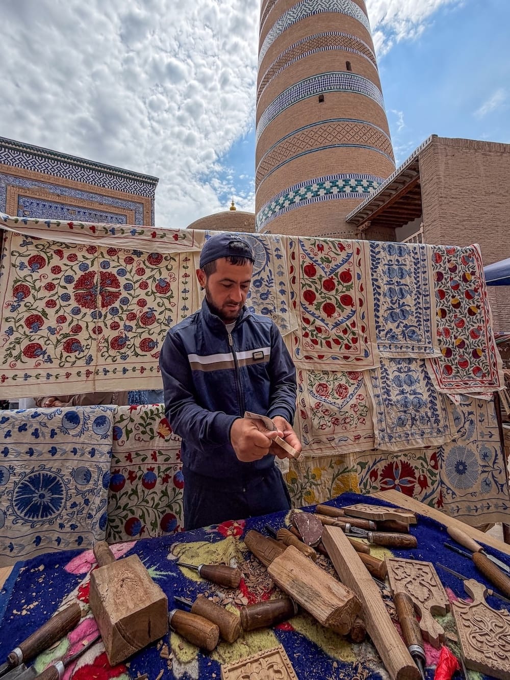 a man standing in front of a table of wood carvings in front of suzanis in Khiva