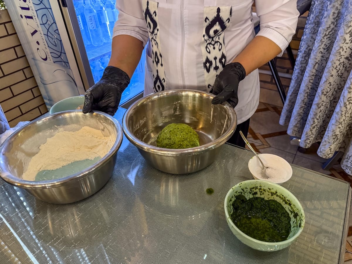 a woman standing behind a bowl of green dill noodles with gloves on