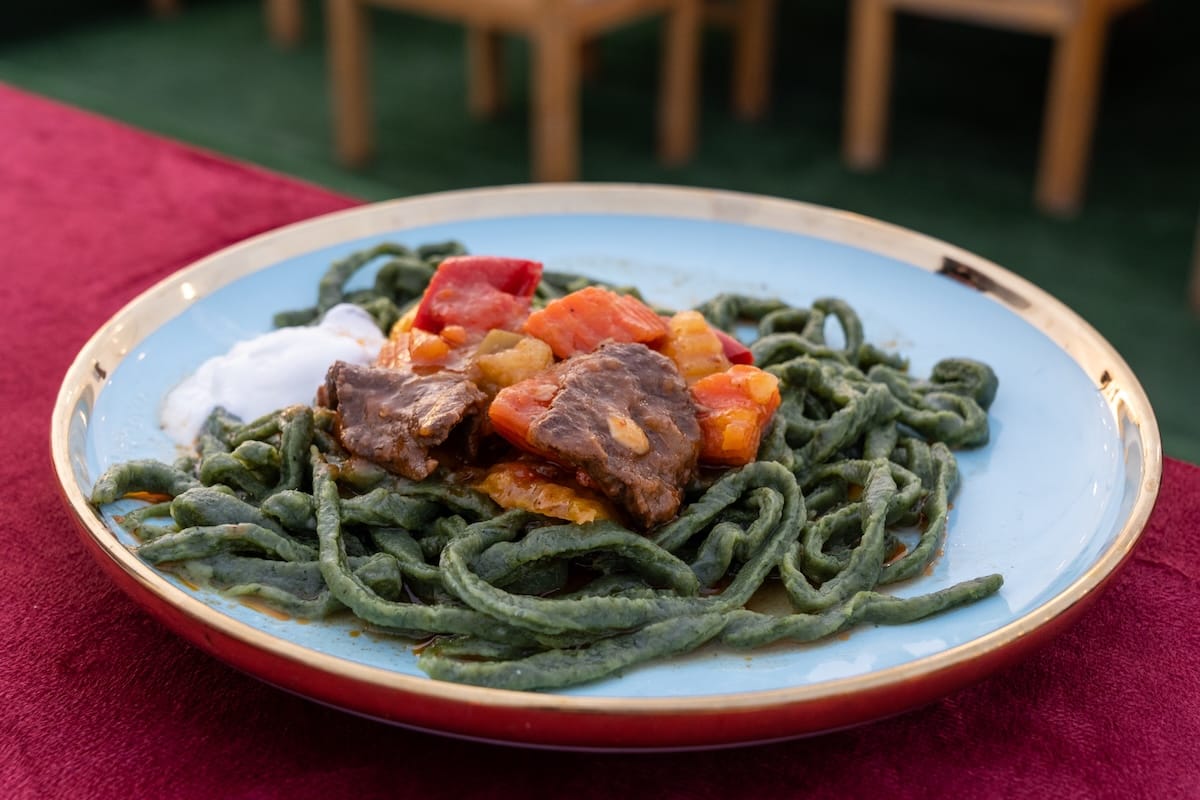 a plate of green noodles with root vegetables, meat, and a yoghurt