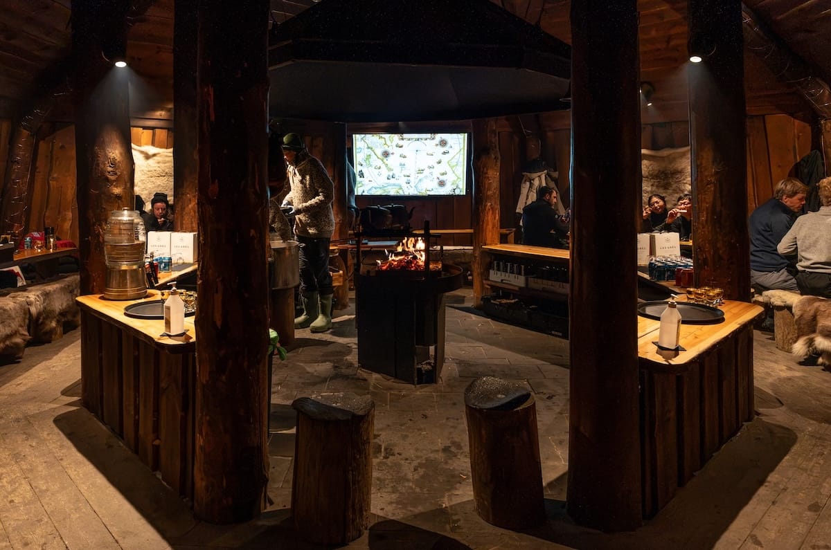 a wooden hut that is kind of dark with a fire in the middle and people sitting at tables around it
