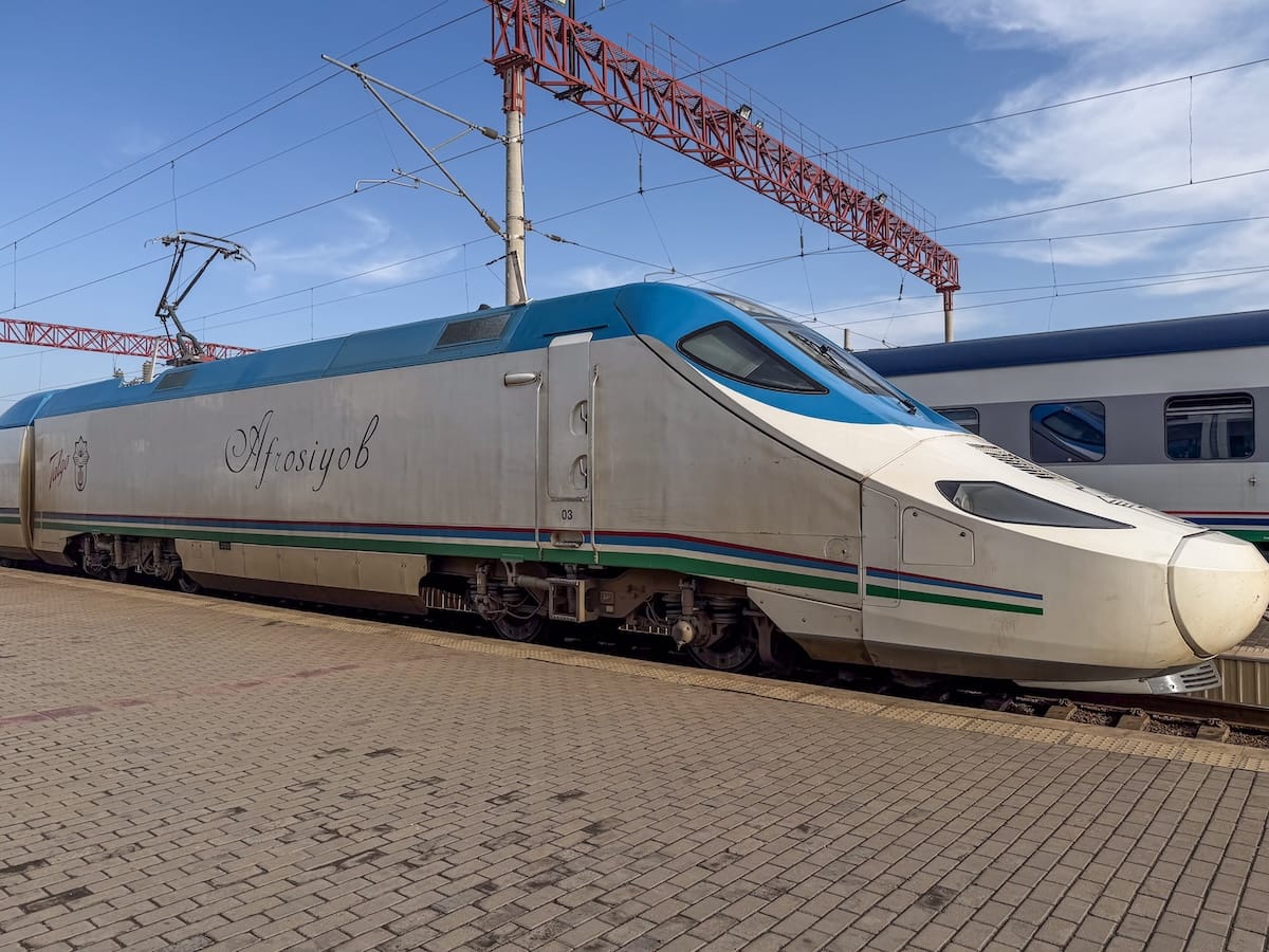 Front of the Afrosiyob high-speed train standing at a station platform, with a silver body, blue roof and green and blue stripes, beneath overhead power lines and a red gantry against a blue sky.