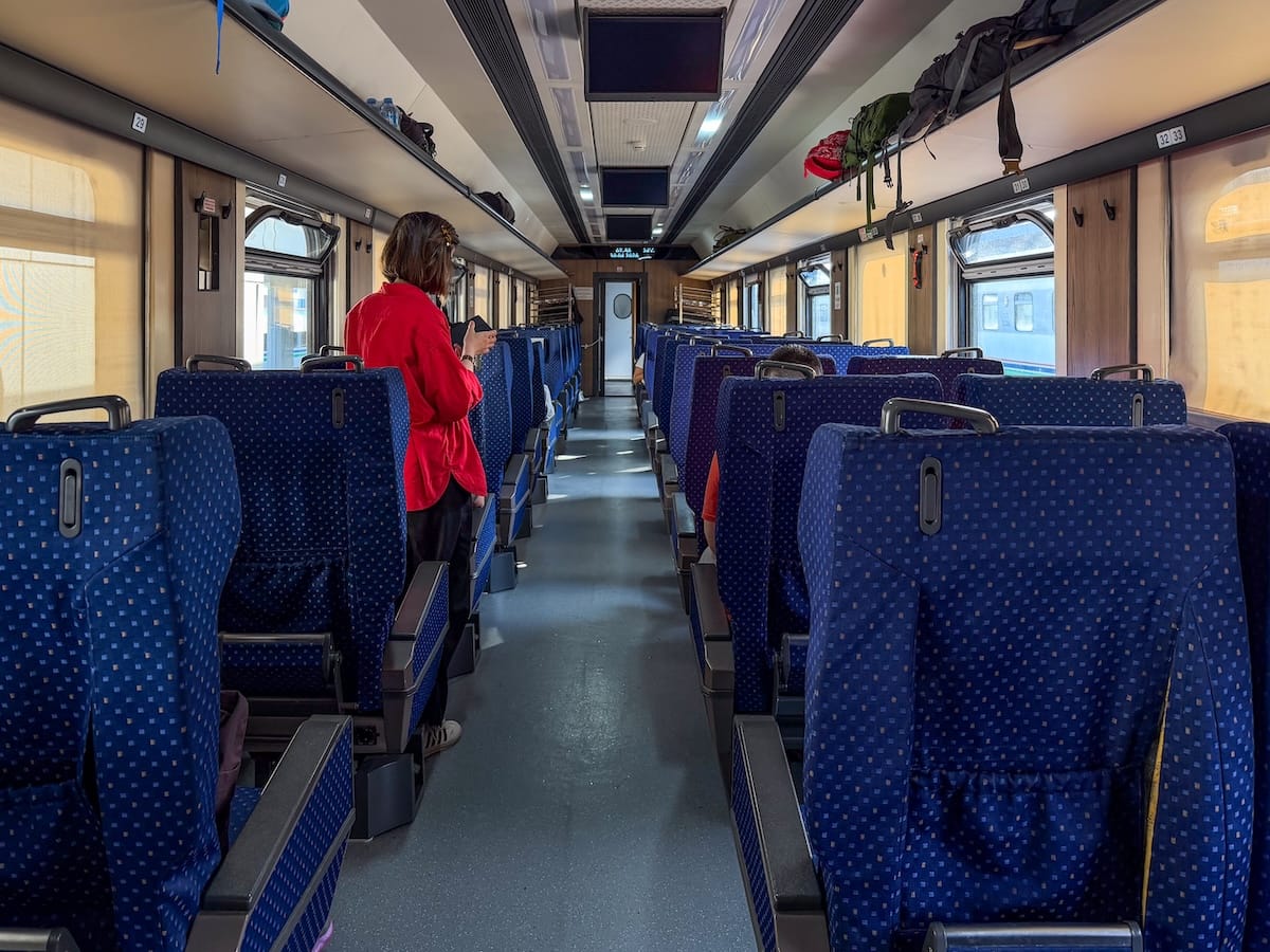 Interior of a train carriage with rows of blue patterned seats on both sides of a central aisle, overhead luggage racks holding backpacks, and a passenger in a red jacket standing beside a seat as sunlight streams through the windows.