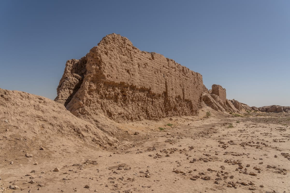 Weathered mud-brick walls of a ruined desert fortress rising above dry, cracked ground scattered with stones, under a clear blue sky in Uzbekistan.