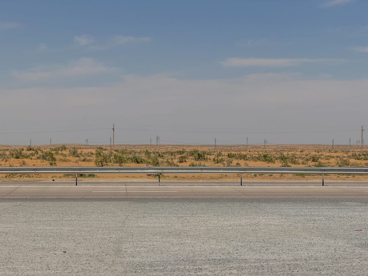 Flat desert landscape of orange sand and low green scrub seen across an empty highway and metal guardrail, with power lines stretching toward a hazy horizon under a pale blue sky.