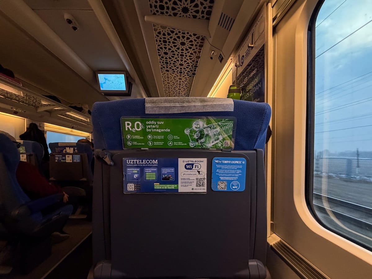 Back of a blue train seat with a folded tray table, stickers for onboard Uztelecom Wi-Fi and contactless NFC payment, and a green water advert, beside a window showing power lines and the track at dusk.